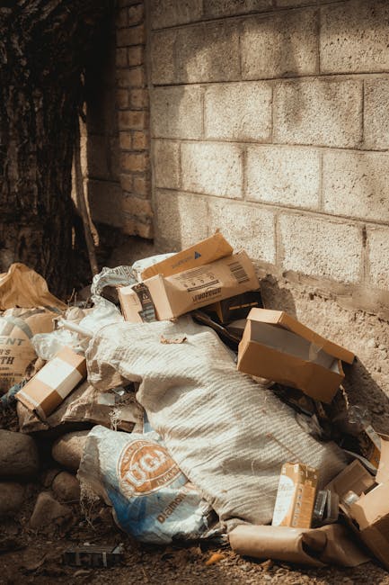 A worker in a red and yellow uniform is operating a large red rubbish collection vehicle positioned on a paved roadside area with a small amount of debris scattered on the ground. The back of the vehi