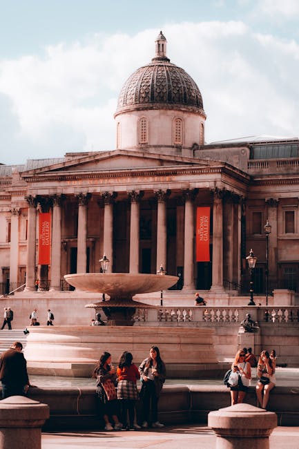 A black and white photograph of Trafalgar Square in Central London, featuring a large, ornate fountain as the central object. The fountain's basin is circular, made of stone with a smooth, polished su