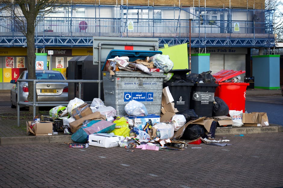 A large collection of mixed waste and rubbish bags, cardboard boxes, and paper or plastic packaging scattered on the pavement near a recycling bin, with a parked grey car located to the left. The recy
