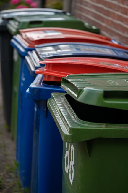 A large collection of mixed waste and rubbish bags, cardboard boxes, and paper or plastic packaging scattered on the pavement near a recycling bin, with a parked grey car located to the left. The recy